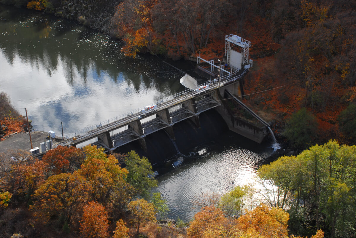 Image of a large dam on a river, with trees changing color on the banks. 