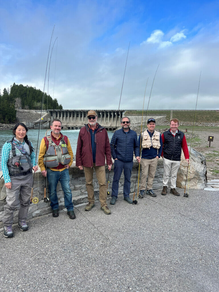 Six smiling people with rods pose in front of the Jackson Lake Dam